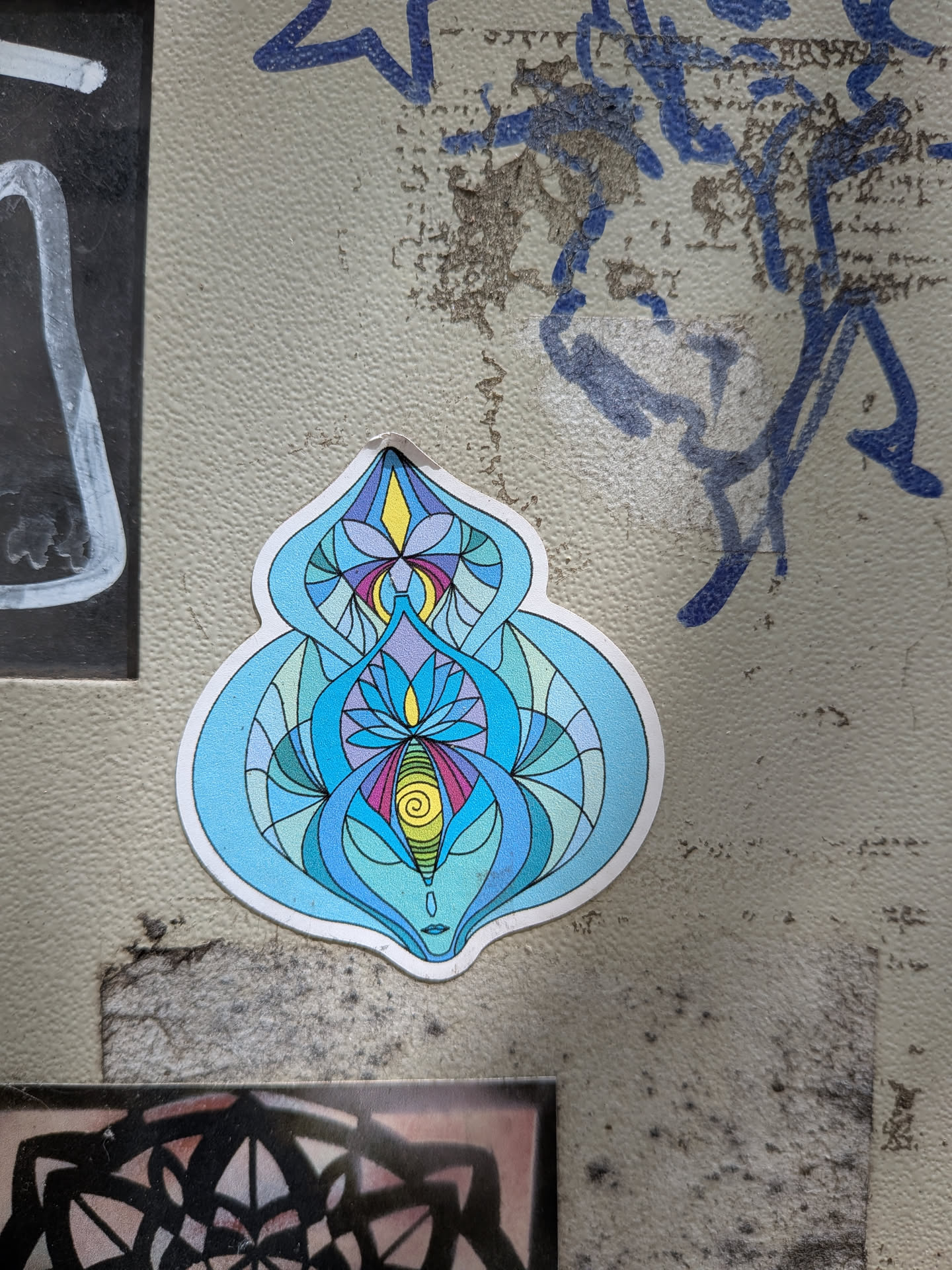 A die-cut sticker featuring a symmetrical, abstract design in shades of blue, teal, purple, and yellow. The intricate linework suggests a stylized lotus, stained glass, or spiritual yoni art, complete with a small yellow spiral at the lower center.