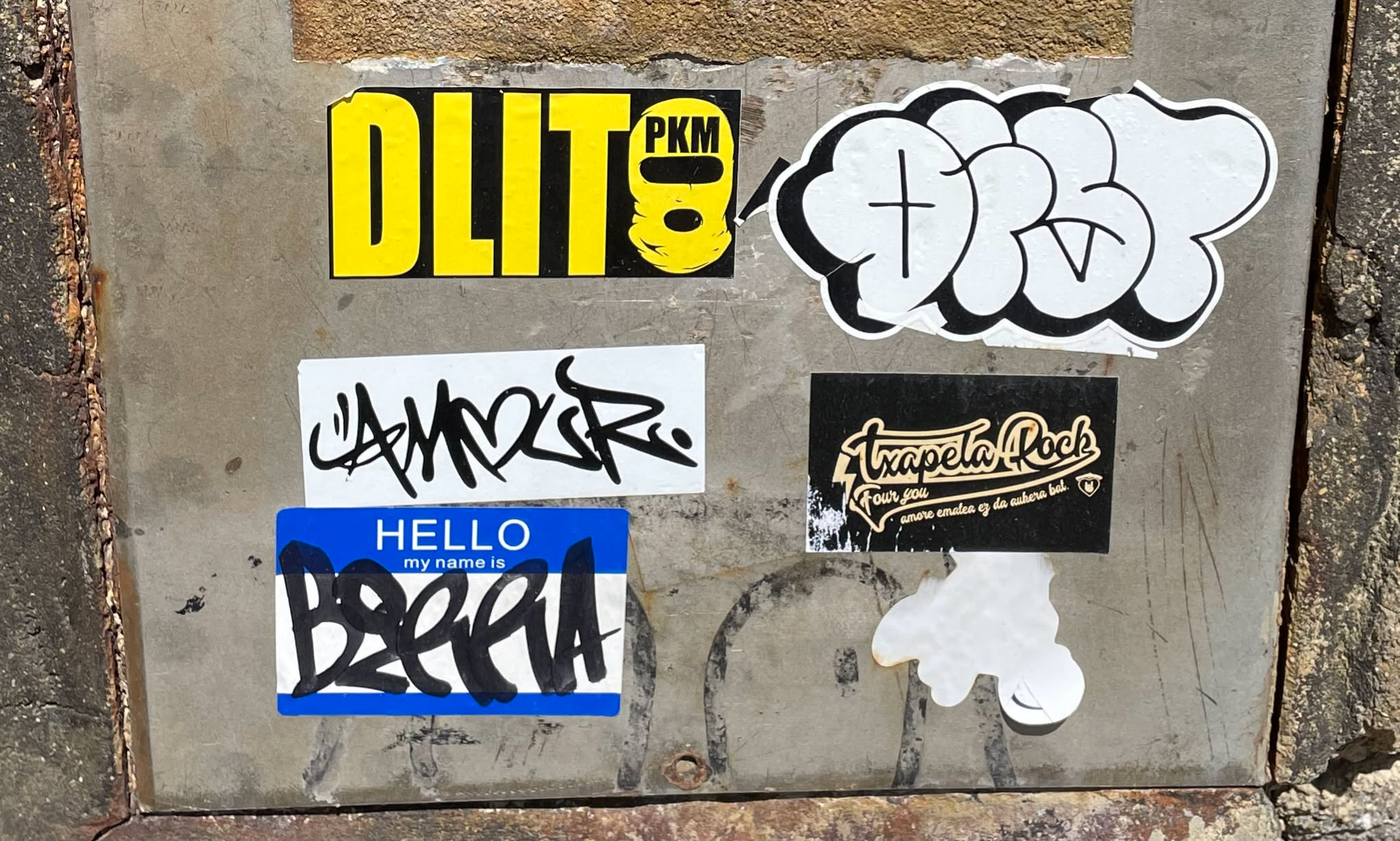 The central sticker is a rectangular, bright yellow design with bold black text. The main text, 'DLIT', is prominently displayed in a sans-serif font. To the right of 'DLIT', within the same yellow sticker, is a circular element containing the black letters 'PKM' above a stylized black icon resembling a face, mask, or helmet. The sticker has clean edges and appears relatively new compared to some surrounding elements, though it shows minor signs of outdoor exposure.