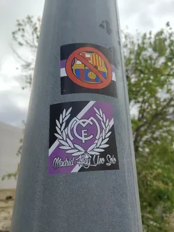 Street sticker The sticker prominently displays a stylized white Real Madrid crest, featuring the intertwined letters 'M' and 'F' topped with a crown, all encased within a classic white laurel wreath. The background is dynamically split by a diagonal line, with one half in solid black and the other in a rich purple. Below the emblem, the phrase 'Madrid Hay Uno Solo' is written in an elegant, flowing white script font. The design is crisp and graphic, conveying strong club loyalty.