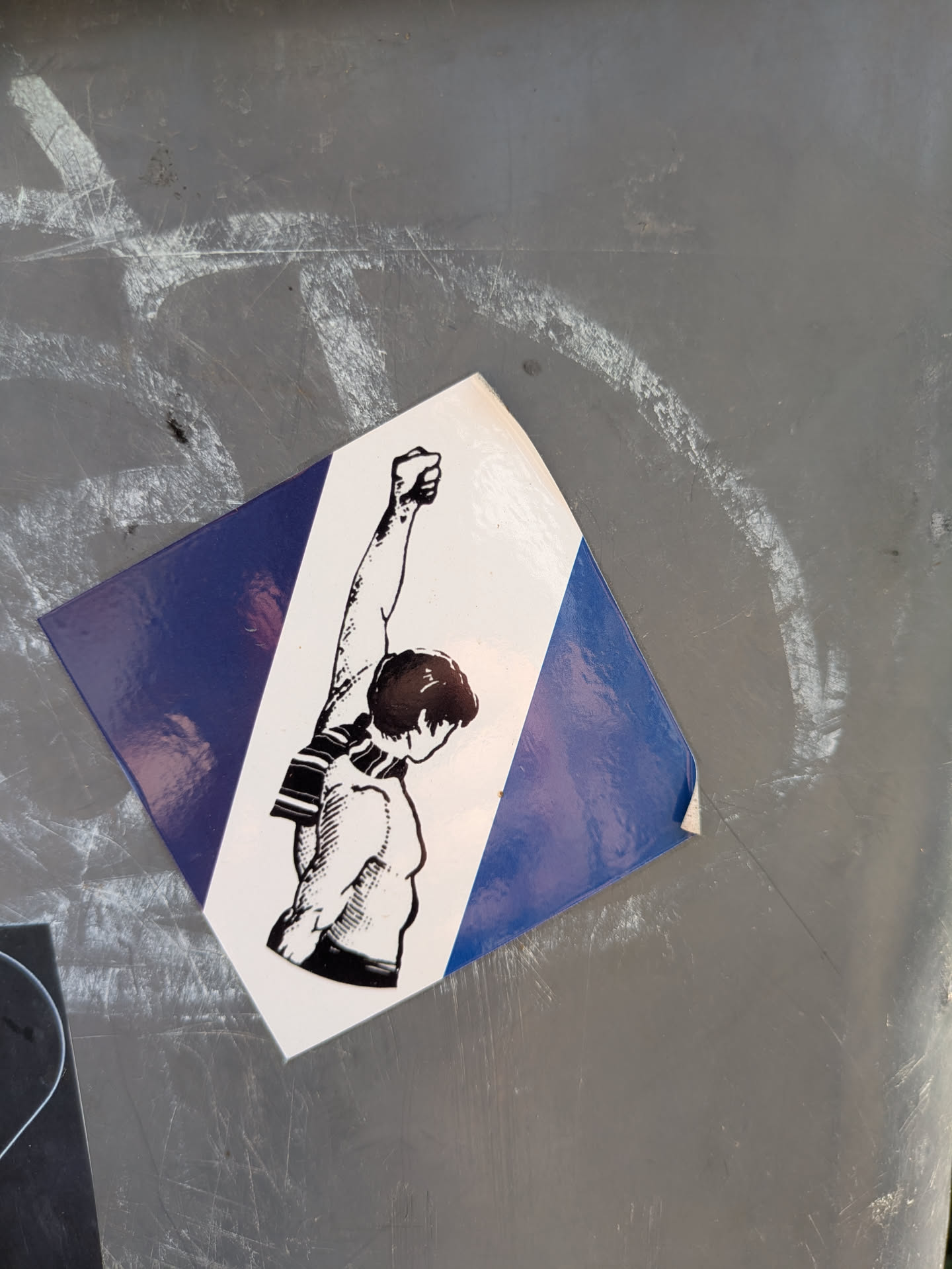 The sticker shows a person with a raised fist, wearing a scarf. The sticker's background is split diagonally, with one section blue and the other white. It's stuck to a gray surface.