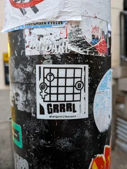 Street sticker A rectangular, black and white sticker featuring a grid pattern resembling a guitar fretboard or musical staff. The grid contains three distinct symbols: a female gender symbol with an '@' sign inside its circle in the top-left, a 'T' in a circle in the top-middle, and a 'B' in a circle in the top-right. Below the grid, the word 'GRRRL' is prominently displayed in bold, uppercase letters. Beneath 'GRRRL', a smaller text reads '@tabgrrrltheband'. The sticker has a white background with black lines and text, showing some minor wear and tear on its edges.