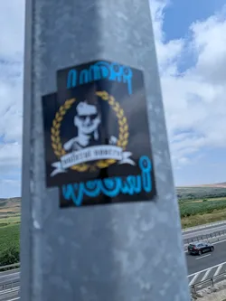 Street sticker Black rectangular sticker featuring a central portrait of a man in sunglasses inside a golden laurel wreath with a white banner. Stylized blue text is positioned at the top and bottom.