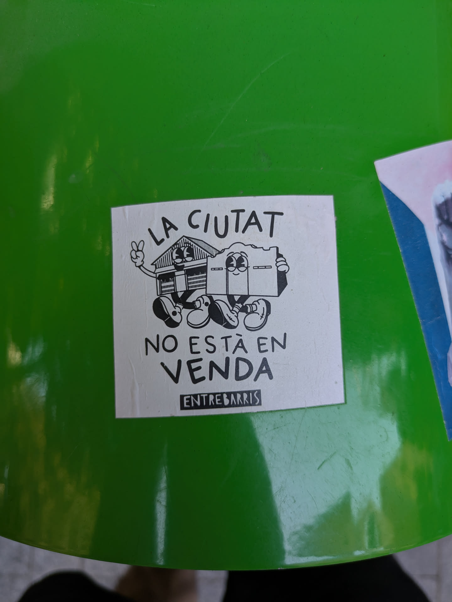 This sticker features a cartoon drawing of a house character with a peace sign giving hand and a box character that holds a sign and wearing shoes. The text on the sticker reads "LA CIUTAT NO ESTA EN VENDA" and "ENTREBARRIS". It seems to be against selling the city. The image is on a green surface.