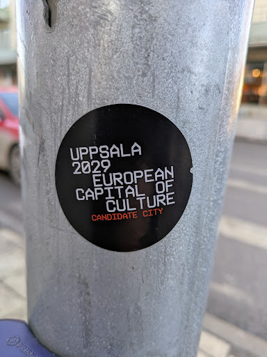 A round, black sticker affixed to a metal pole advertises Uppsala as a candidate city to become the European Capital of Culture in 2029. The text is in white, except for "CANDIDATE CITY" which is in orange.