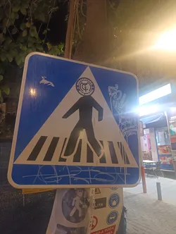 Street sticker The sticker is applied to a standard blue and white pedestrian crossing sign. It features a central black silhouette of a walking person, which is part of the original sign. Above this figure, a circular white logo is visible, containing the text 'BERSERKERS' at the top, 'REAL MADRID' at the bottom, and the Real Madrid crest in the center, flanked by '20' and '05'. To the left of the walking figure, in the blue section of the sign, there's a white, abstract, somewhat bird-like or stylized shape. To the right, a white outline of a hand making a V-sign or peace sign is depicted, with a dripping or melting effect. At the very bottom of the sticker, over the white stripes of the pedestrian crossing, there is white, stylized graffiti or tagging. The sticker appears to be a white vinyl overlay, allowing the blue and white of the original sign to serve as background colors for the added white and black elements.