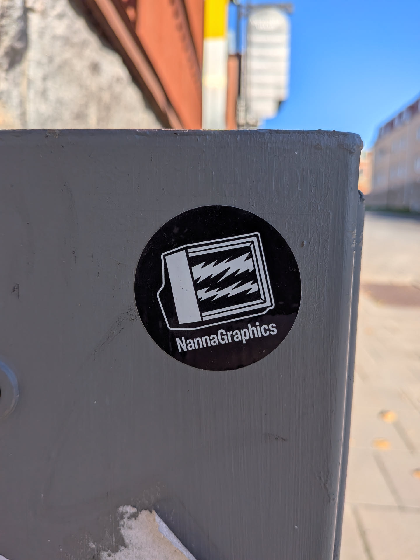 The sticker is circular with a black background. In the center, there is a white graphic that appears to be a stylized representation of a screen or a window with jagged lines inside, suggesting energy or data. Below the graphic, the text 'NannaGraphics' is written in white, sans-serif capital letters. The overall design is clean and modern.