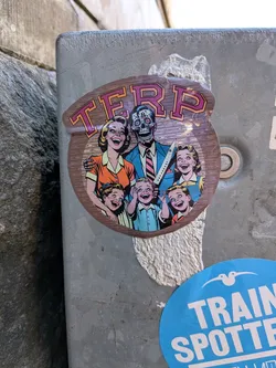 Street sticker The sticker is oval-shaped with a wooden border. It depicts a stylized, retro illustration of a family. The father, in the center, has a skull for a face with exposed teeth and red eyes, wearing a suit and tie and holding a large knife. The mother, to his left, is smiling broadly, wearing an orange shirt. Two children are in the foreground, also smiling widely. Another child is behind the mother. The background has a wood grain texture. The word 'TERP' is prominently displayed in large, stylized letters at the top of the sticker.