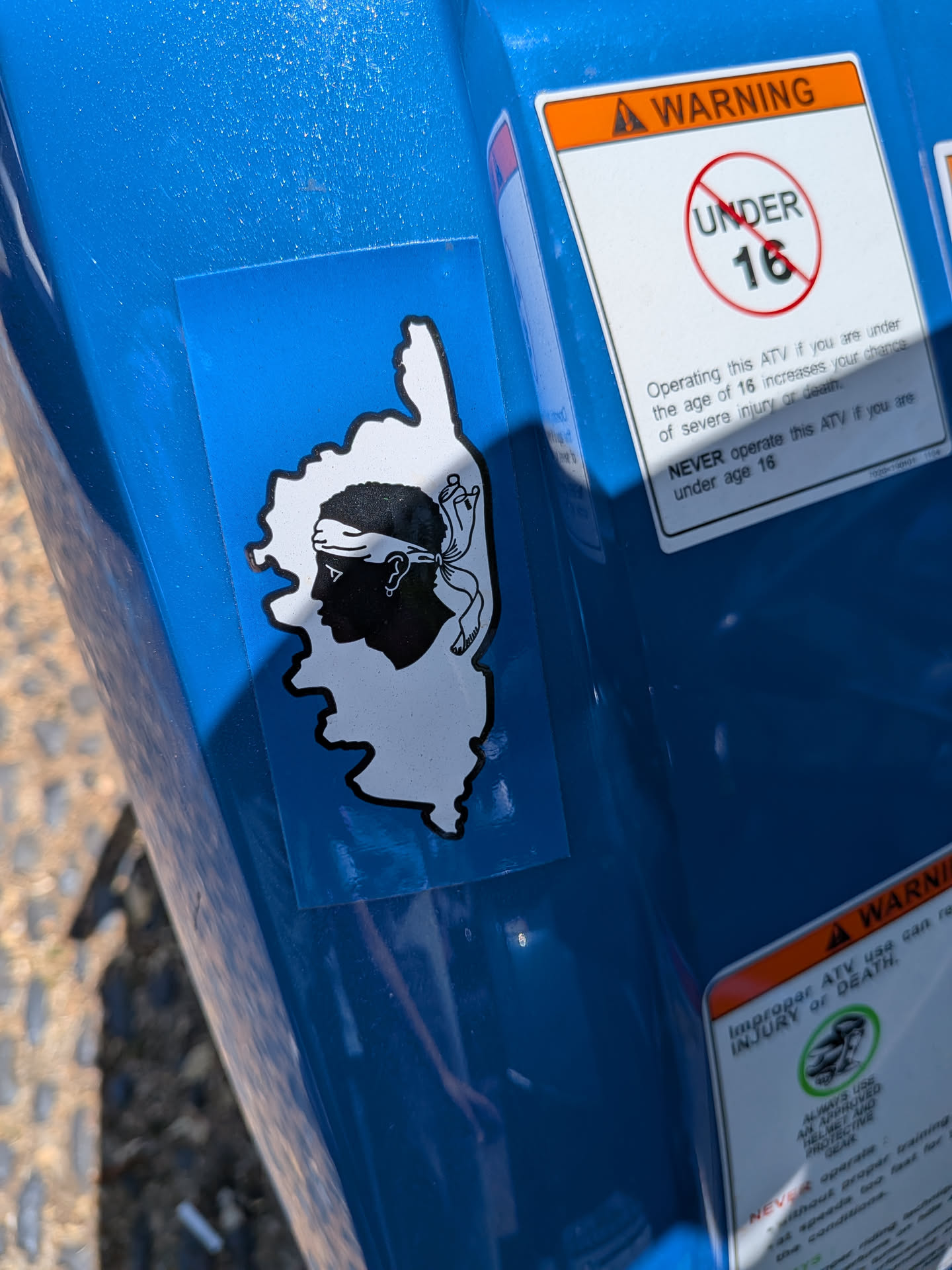 Rectangular blue sticker featuring a white silhouette of Corsica containing the black Moor's Head symbol with a white bandana.