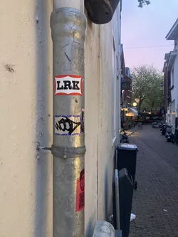 Street sticker The sticker is prominently displayed at the center of the image, adhered to a metallic pipe. It features the letters 'LRK' in bold, sans-serif font, colored black. The text is set against a white background with a red border, creating a strong contrast. The sticker has a slightly worn appearance, suggesting it has been exposed to the elements.