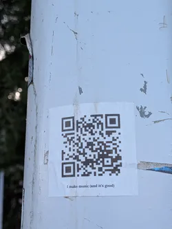 A square white sticker featuring a black QR code with the text 'I make music (and it's good)' printed below, affixed to a weathered white post.