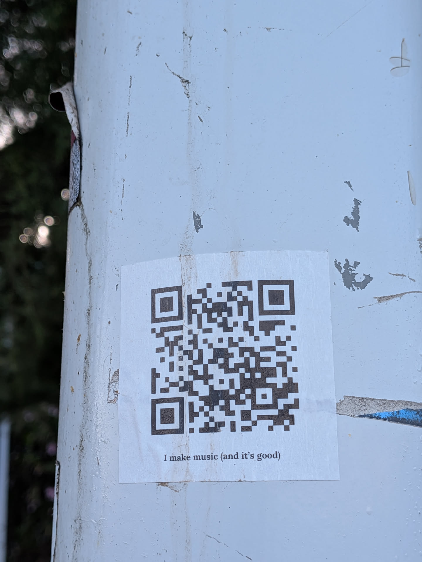 A square white sticker featuring a black QR code with the text 'I make music (and it's good)' printed below, affixed to a weathered white post.