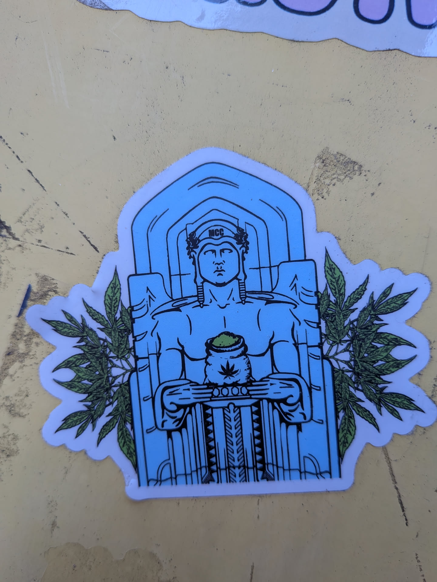 The sticker features a central figure resembling a classical statue, rendered in blue and black lines. The figure is depicted with a stern expression, wearing a laurel wreath on its head with the letters 'MCC' inscribed on it. It holds a sack filled with green material, from which a cannabis leaf symbol is visible. The figure is framed by an architectural element resembling a temple or archway. On either side of the figure, stylized green marijuana leaves with detailed venation extend outwards, creating a symmetrical design. The sticker has a white border and appears to be die-cut to the shape of the illustration.