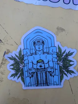 Street sticker The sticker features a central figure resembling a classical statue, rendered in blue and black lines. The figure is depicted with a stern expression, wearing a laurel wreath on its head with the letters 'MCC' inscribed on it. It holds a sack filled with green material, from which a cannabis leaf symbol is visible. The figure is framed by an architectural element resembling a temple or archway. On either side of the figure, stylized green marijuana leaves with detailed venation extend outwards, creating a symmetrical design. The sticker has a white border and appears to be die-cut to the shape of the illustration.