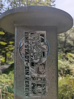 Street sticker The sticker is rectangular and vertically aligned on a grey metal pole. It features two main vertical text blocks. On the left, in a more straightforward blocky font, it reads "EINE STADT - EIN VEREIN". On the right, the word "RAGAZZA" is displayed in a stylized, somewhat graffiti-inspired font with ornate details and patterns filling the letters. The background of the sticker appears to be a mix of grey and white with subtle patterns, possibly illustrating the club's crest or symbolic elements. A small circular blue and white sticker with a cartoon character (possibly a sailor or a figure with an anchor tattoo) is partially visible behind the main sticker on the left side, but it is largely obscured. The setting is outdoors, with blurred green foliage and trees in the background under a bright sky.