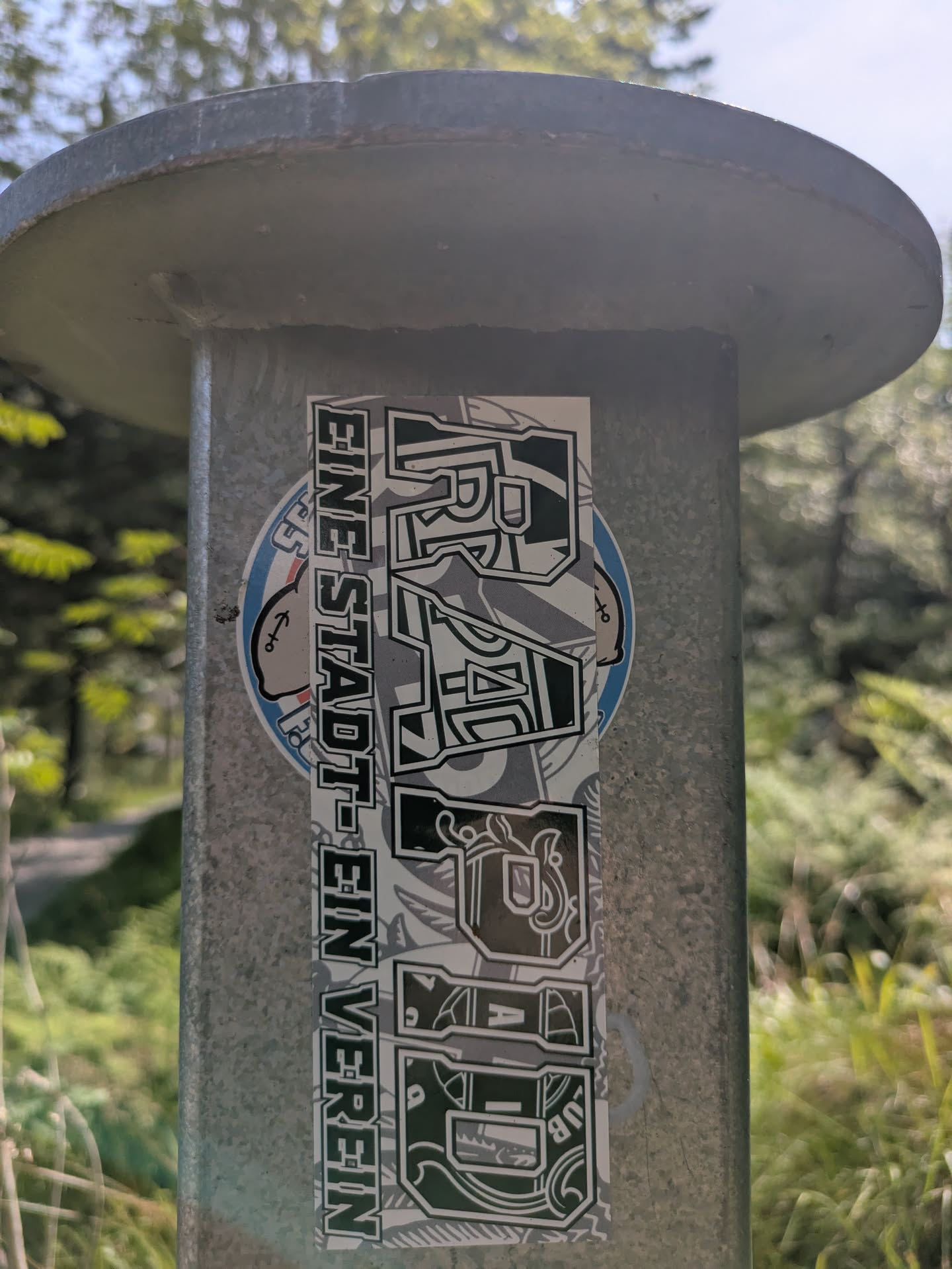 The sticker is rectangular and vertically aligned on a grey metal pole. It features two main vertical text blocks. On the left, in a more straightforward blocky font, it reads "EINE STADT - EIN VEREIN". On the right, the word "RAGAZZA" is displayed in a stylized, somewhat graffiti-inspired font with ornate details and patterns filling the letters. The background of the sticker appears to be a mix of grey and white with subtle patterns, possibly illustrating the club's crest or symbolic elements. A small circular blue and white sticker with a cartoon character (possibly a sailor or a figure with an anchor tattoo) is partially visible behind the main sticker on the left side, but it is largely obscured. The setting is outdoors, with blurred green foliage and trees in the background under a bright sky.