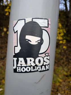 Street sticker The sticker is centrally located on a light gray pole. It features a stylized illustration of a person wearing a black balaclava that covers their head and neck, leaving only their eyes visible. The eyes are depicted with white irises and black pupils, giving a stern or intense look. To the left and right of the head are large white numbers, '1' and '5', outlined in black. The background behind the head and numbers has a subtle pinkish hue. Below the masked figure, the word 'JAROŠ' is prominently displayed in large, bold, white capital letters with a black outline. Beneath 'JAROŠ', the word 'HOOLIGAN' is written in smaller, white capital letters, also with a black outline. The sticker appears to be made of paper and shows some signs of wear, with creases and slightly peeling edges.