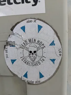 Street sticker A circular sticker features the words "DEAD MEN HOPS" in bold lettering around a skull with crossed hop bines. Text "ESTABLISHED '70'7" is at the bottom. The sticker's edges are worn and torn.