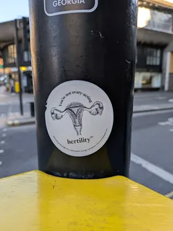 Street sticker A circular, off-white sticker featuring a detailed black and white line drawing of a human uterus with fallopian tubes and ovaries at its center. Above the illustration, text curves along the top edge reading "You're not ovary-acting". Below the illustration, the word "hertility®" is prominently displayed. Along the bottom curve of the sticker, smaller text reads "Specialist gynaecology from menstruation through menopause. www.hertilityhealth.com". The sticker has a slightly worn appearance, with some minor scuffs and a small tear on its left edge.