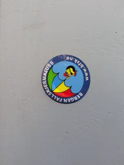 Street sticker A circular sticker predominantly blue, with a lighter blue central area. The outer blue ring contains white text. The center features a stylized cartoon character, appearing to be a person, beneath a multi-colored parachute canopy (segments of green, yellow, and red). The text 'BERGEN FALLSKJERMKLUBB' is written along the bottom curve, and 'www.bfsk.no' is written along the top curve.