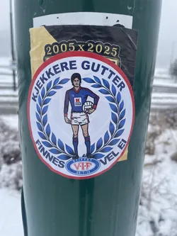 Street sticker The central sticker is circular, featuring a stylized male football player in a retro blue and red kit, holding a white football. He stands within a white background, framed by a blue laurel wreath. Around the outer edge, on a red band, blue text reads "KJEKKERE GUTTER" at the top and "FINNES VEL EI" at the bottom. Below the player, a small blue oval crest contains "STIFTET V.I.F 29-7-13" in white. The player's shirt displays "UMBRO", a 'K' logo, and "KASKO". Partially visible behind this main sticker is a rectangular one with a black background and gold text "2005 x 2025". The stickers are affixed to a dark green pole, with a snowy, foggy background.