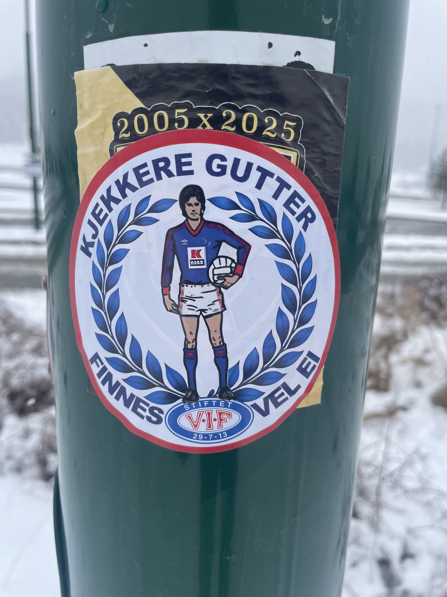 The central sticker is circular, featuring a stylized male football player in a retro blue and red kit, holding a white football. He stands within a white background, framed by a blue laurel wreath. Around the outer edge, on a red band, blue text reads "KJEKKERE GUTTER" at the top and "FINNES VEL EI" at the bottom. Below the player, a small blue oval crest contains "STIFTET V.I.F 29-7-13" in white. The player's shirt displays "UMBRO", a 'K' logo, and "KASKO". Partially visible behind this main sticker is a rectangular one with a black background and gold text "2005 x 2025". The stickers are affixed to a dark green pole, with a snowy, foggy background.