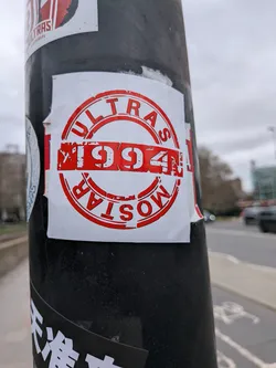 Street sticker A square-shaped sticker with a white background, featuring a prominent circular design in red. The word 'ULTRAS' is written in capital letters along the top curve of the circle, while 'MOSTAR' is written inverted along the bottom curve. In the center, the year '1994' is displayed horizontally in large, distressed red characters. The red elements have a textured, slightly worn appearance, giving the sticker a grunge or vintage feel. The sticker is slightly peeling at the edges, revealing its adhesive nature.