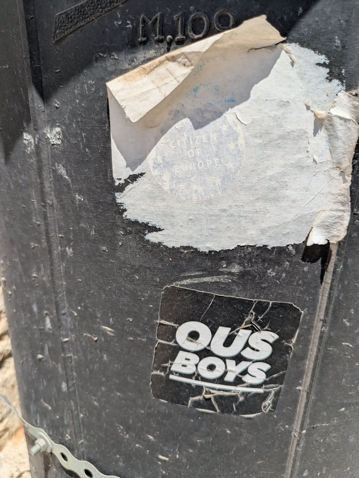 Close-up of a worn sticker with the text "OUS BOYS" in bold white letters. The sticker is on a black, textured surface, and the sticker itself is cracked and aged.