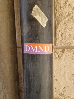 A close-up shot of a DMND. sticker affixed to a dark-colored pipe. The sticker is rectangular with an orange background and purple text.  The pipe is weathered and shows signs of wear.