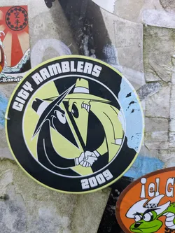 Street sticker The central sticker is a circular design with a thick black outer ring and a vibrant yellow inner circle. The primary imagery consists of two stylized, long-nosed cartoon characters in fedora-like hats, one entirely black and the other primarily yellow/lime green, facing each other and shaking hands. Their design strongly echoes the 'Spy vs. Spy' characters from Mad Magazine. The text 'CITY RAMBLERS' is printed in white block letters along the top arc of the black outer ring, and '2009' is at the bottom. A distressed light blue patch is visible on the right edge of the black outer ring.