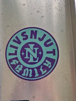 Street sticker Close-up of a round sticker with the text "LIVSNJUT" circling a central logo featuring the letters "NL" within a stylized emblem. The sticker has a teal and purple color scheme and appears to be affixed to a metallic surface.