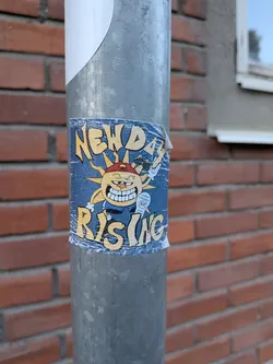 Street sticker The sticker features a cartoonish sun character with a wide, toothy grin and a red baseball cap worn backward. The sun has spiky yellow rays emanating from its head and is depicted with arms and legs. It's wearing a blue shirt and appears to be holding a white object, possibly a microphone or a spray can, with a small circular emblem labeled 'NDR' on it. The background of the sticker is a dark blue with a textured, almost distressed appearance. The text 'NEW DAY' is prominently displayed in yellow, stylized lettering at the top, and 'RISING' in similar lettering at the bottom. The sticker shows signs of wear and tear, with some peeling at the edges, suggesting it has been applied outdoors.