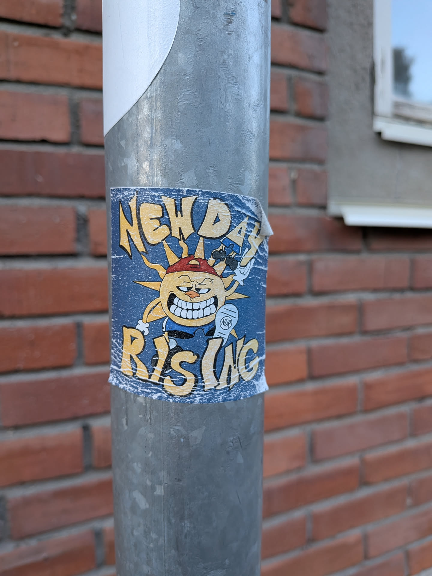 The sticker features a cartoonish sun character with a wide, toothy grin and a red baseball cap worn backward. The sun has spiky yellow rays emanating from its head and is depicted with arms and legs. It's wearing a blue shirt and appears to be holding a white object, possibly a microphone or a spray can, with a small circular emblem labeled 'NDR' on it. The background of the sticker is a dark blue with a textured, almost distressed appearance. The text 'NEW DAY' is prominently displayed in yellow, stylized lettering at the top, and 'RISING' in similar lettering at the bottom. The sticker shows signs of wear and tear, with some peeling at the edges, suggesting it has been applied outdoors.