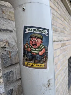 Street sticker The sticker features a cartoon illustration of an angry, red-faced baby wearing a green flat cap and a green and white striped shirt. The baby is sitting down, holding a bottle of beer in its right hand and looking furious. There are other beer bottles scattered around its feet, some of which appear to be empty or knocked over. The background is a dark blue, textured pattern. Above the character, a banner reads 'VSK GREEN KIDS' in white text with a green outline. Below the character, a yellow oval shape contains the text 'VSK OLD TIMERS' in black lettering. The overall style is reminiscent of vintage cartoons or comic art.