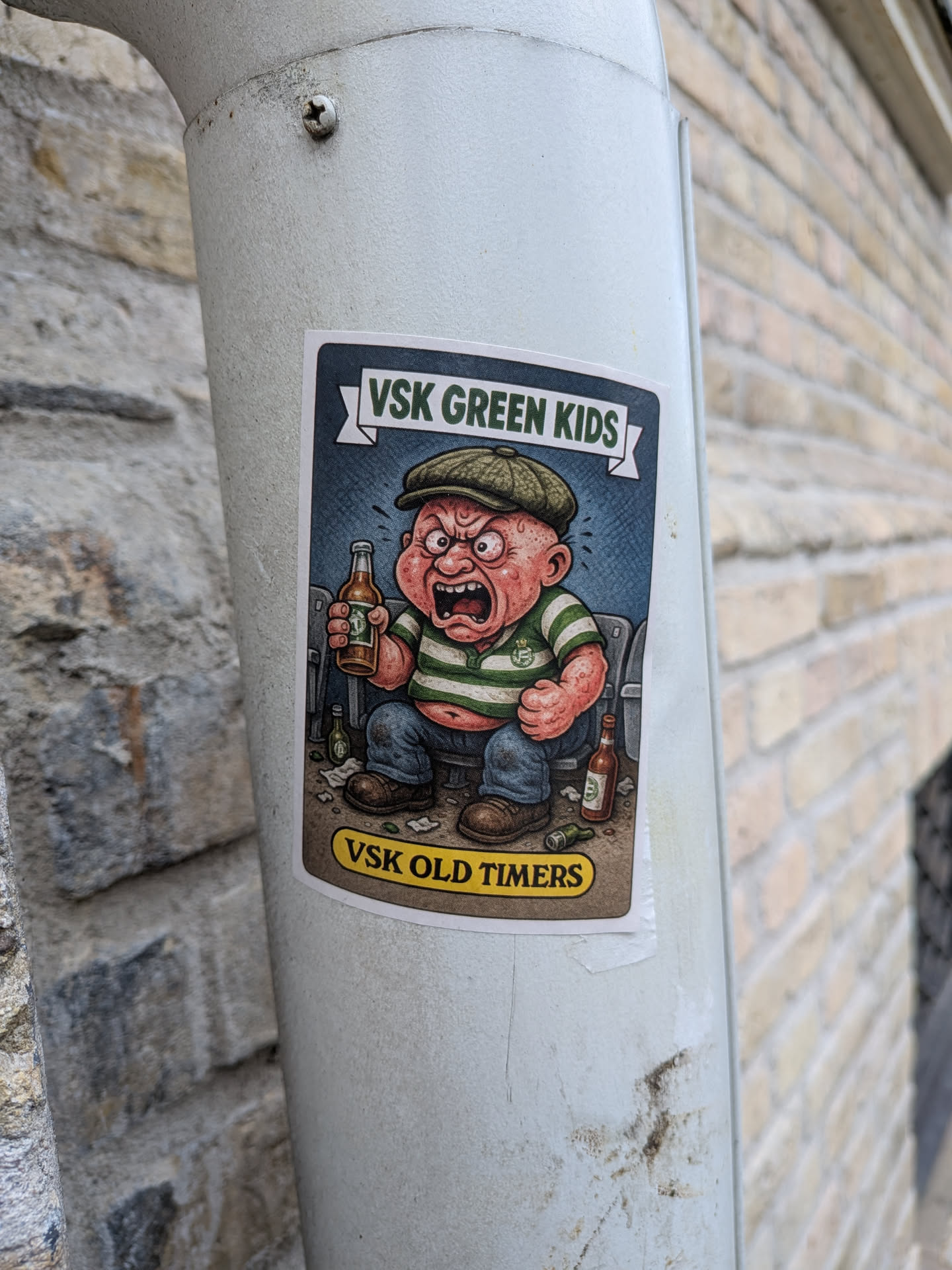 The sticker features a cartoon illustration of an angry, red-faced baby wearing a green flat cap and a green and white striped shirt. The baby is sitting down, holding a bottle of beer in its right hand and looking furious. There are other beer bottles scattered around its feet, some of which appear to be empty or knocked over. The background is a dark blue, textured pattern. Above the character, a banner reads 'VSK GREEN KIDS' in white text with a green outline. Below the character, a yellow oval shape contains the text 'VSK OLD TIMERS' in black lettering. The overall style is reminiscent of vintage cartoons or comic art.