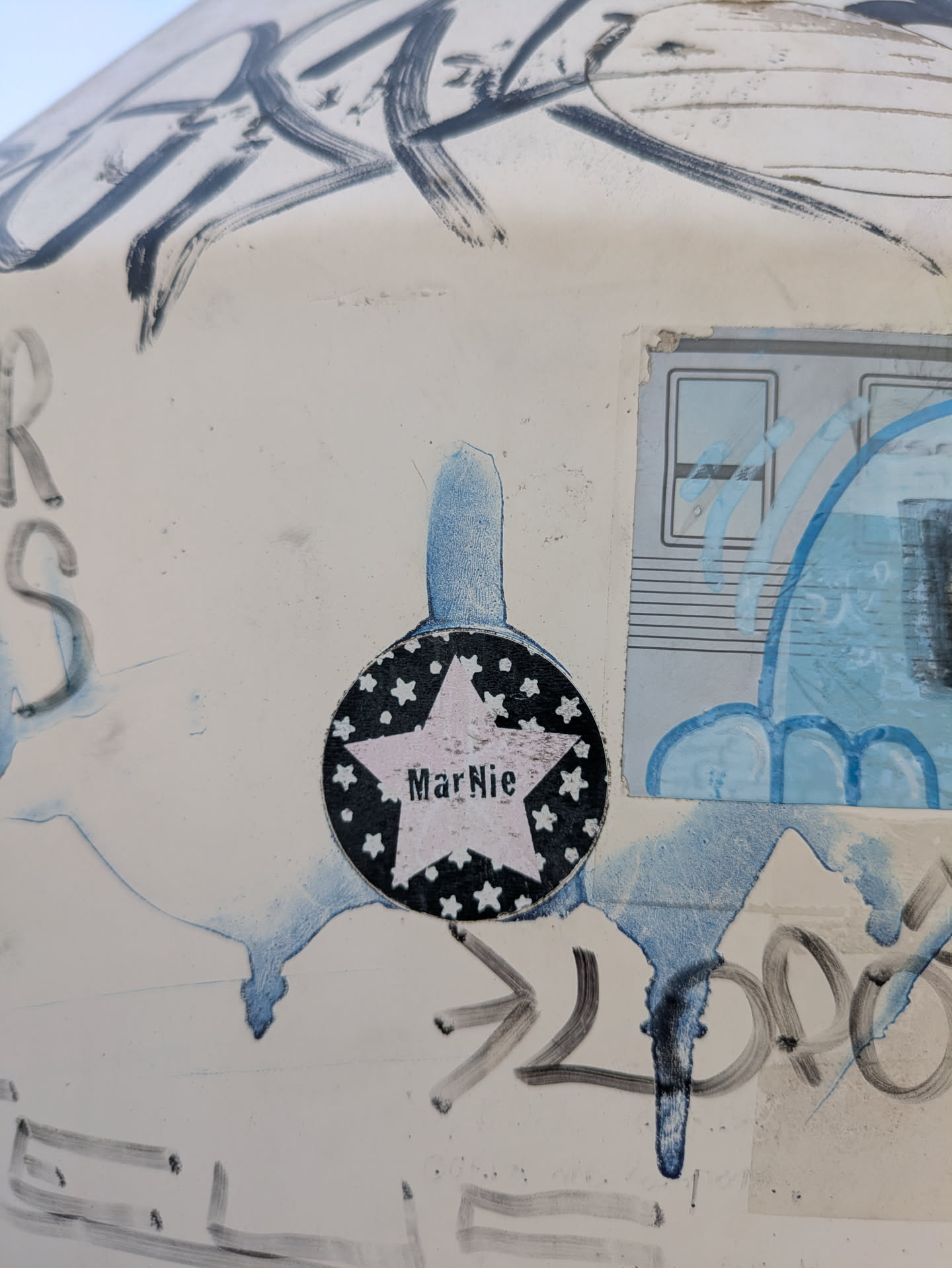 The sticker features a black circle with white stars around the edge. Inside the circle is a star shape with the name "MarNie" printed inside. The sticker looks to be aged and slightly damaged. It is affixed to a surface that also has other markings and stickers on it.
