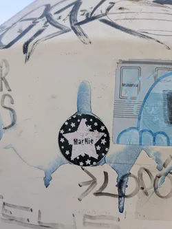 Street sticker The sticker features a black circle with white stars around the edge. Inside the circle is a star shape with the name "MarNie" printed inside. The sticker looks to be aged and slightly damaged. It is affixed to a surface that also has other markings and stickers on it.