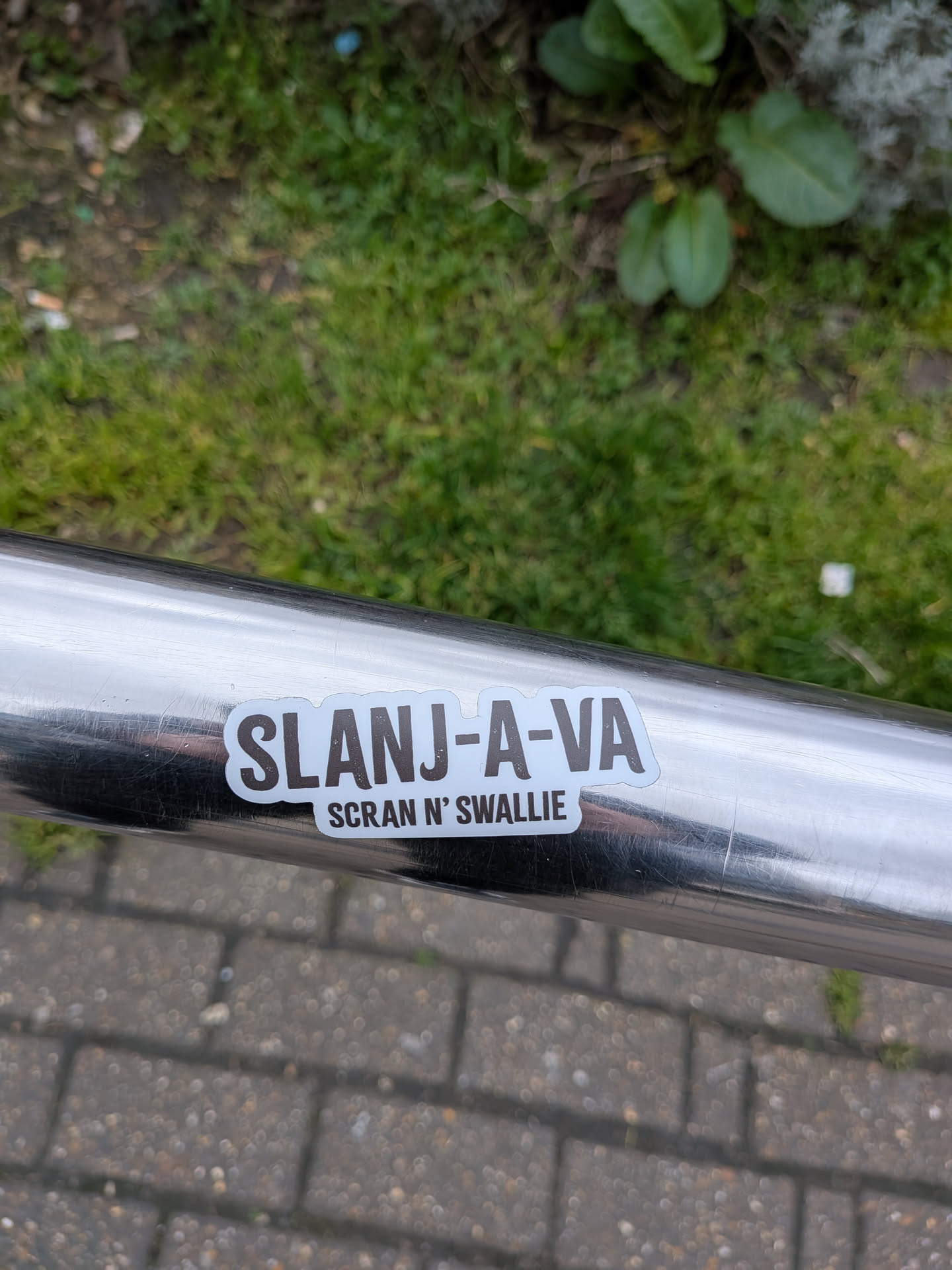 The sticker is rectangular with rounded corners, featuring a white background and a thin black outline. The main text, "SLANJ-A-VA", is prominently displayed in large, bold, dark brown (almost black) sans-serif capital letters. Below it, in smaller, less bold, dark brown capital letters, is the phrase "SCRAN N' SWALLIE". The sticker is applied to a shiny, scratched silver metal railing, with reflections visible on the railing's surface.
