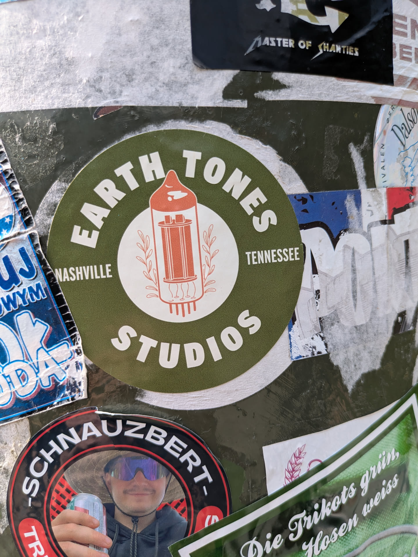A circular sticker with an olive green background and white text, centered around a reddish-orange illustration of a vacuum tube. The text 'EARTH TONES' is at the top, 'STUDIOS' at the bottom, 'NASHVILLE' on the left, and 'TENNESSEE' on the right. The vacuum tube is surrounded by decorative leafy branches, giving it a classic audio or music production aesthetic.
