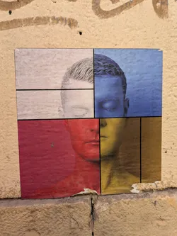 Street sticker The sticker shows a man's face split into four rectangular sections, each in a different color: white, blue, red, and gold. The face has eyes closed. The sticker is adhered to a textured wall.