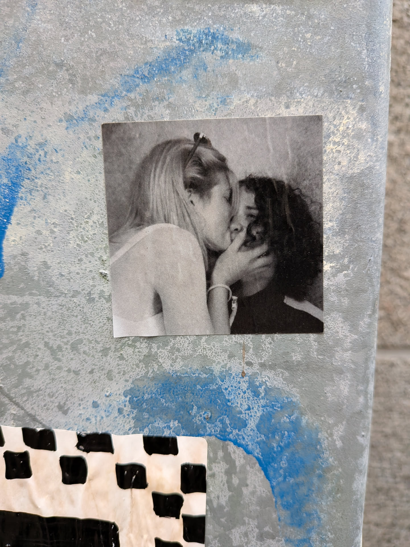 Street sticker The sticker is a black and white photograph featuring two individuals, appearing to be a couple, sharing a kiss. The person on the left has blonde hair styled up and is wearing a light-colored top. Their hand is gently touching the face of the person on the right, who has dark, curly hair and is wearing a dark top. The image has a slightly grainy texture, typical of older photographs or a deliberate artistic choice. The sticker is square-shaped and appears to be adhered to a textured, metallic surface with blue paint splatters.