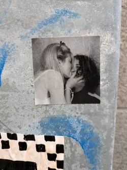 Street sticker The sticker is a black and white photograph featuring two individuals, appearing to be a couple, sharing a kiss. The person on the left has blonde hair styled up and is wearing a light-colored top. Their hand is gently touching the face of the person on the right, who has dark, curly hair and is wearing a dark top. The image has a slightly grainy texture, typical of older photographs or a deliberate artistic choice. The sticker is square-shaped and appears to be adhered to a textured, metallic surface with blue paint splatters.