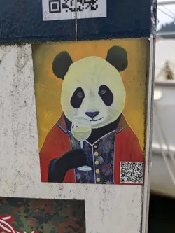 Street sticker The central sticker depicts a panda, stylized and anthropomorphic, wearing a deep red jacket with blue accents and a richly patterned, possibly floral or botanical, dark blue vest. The panda holds a stemmed glass filled with a light-colored liquid, presumably white wine, in its left hand. Its expression is calm and somewhat sophisticated. The background behind the panda is a solid, vibrant yellow-orange. The sticker has a somewhat weathered appearance, suggesting it has been exposed to the elements.