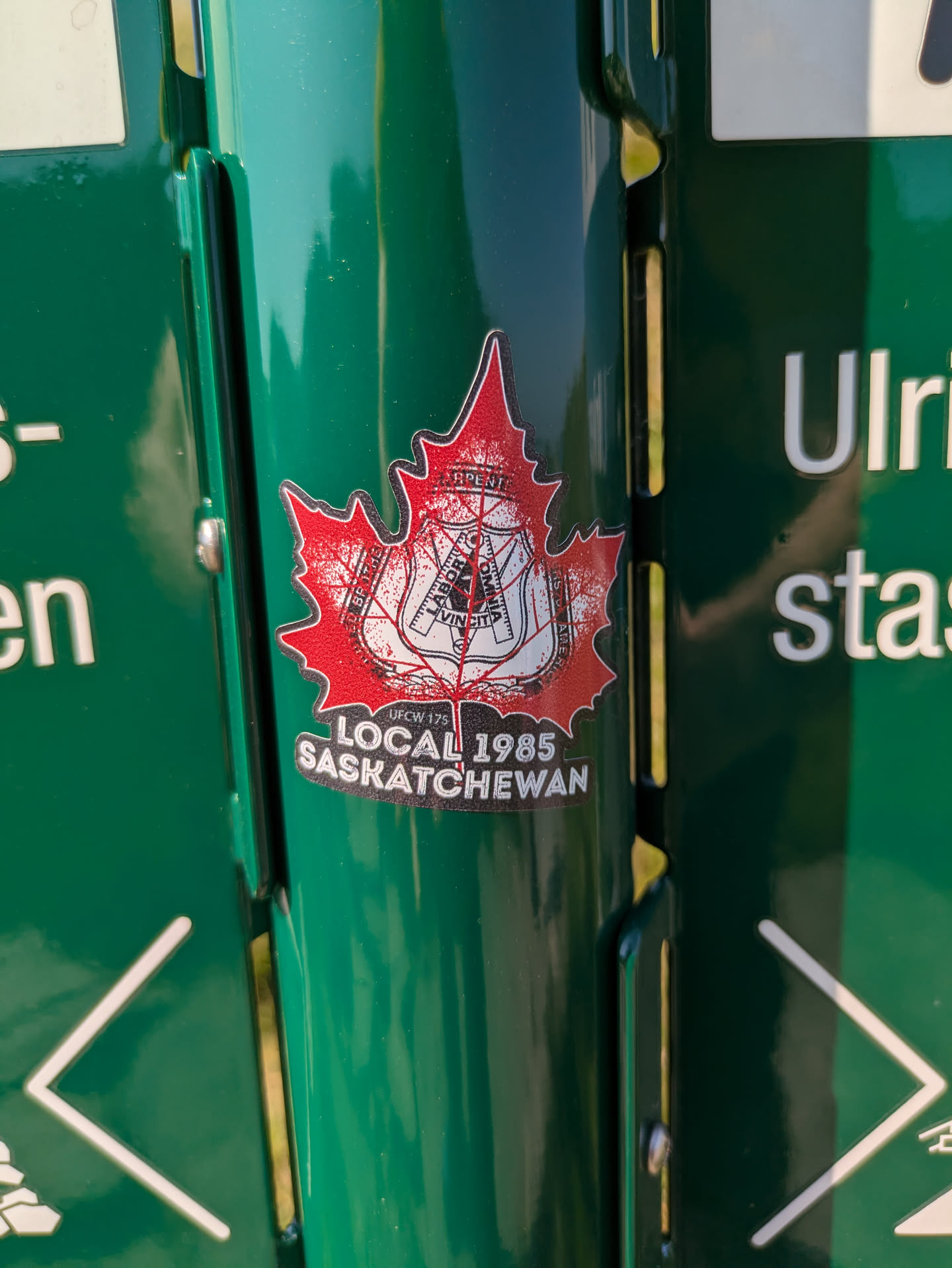 The sticker is shaped like a classic red Canadian maple leaf with a bright white border, affixed to a dark green metallic surface. Prominently displayed within the maple leaf is a black and white emblem belonging to the United Brotherhood of Carpenters and Joiners of America, which includes a shield with traditional tools (compass, square, plane) and the Latin motto "LABOR OMNIA VINCIT" (Labor Conquers All). Surrounding the shield are the words "UNITED BROTHERHOOD OF CARPENTERS AND JOINERS OF AMERICA". Below this central emblem, against the red background of the maple leaf, smaller white text reads "UFCW 175" followed by larger white text stating "LOCAL 1985 SASKATCHEWAN". The red surface of the maple leaf has a slightly textured or speckled appearance.