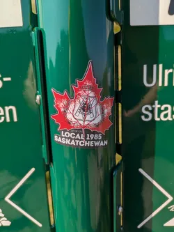 Street sticker The sticker is shaped like a classic red Canadian maple leaf with a bright white border, affixed to a dark green metallic surface. Prominently displayed within the maple leaf is a black and white emblem belonging to the United Brotherhood of Carpenters and Joiners of America, which includes a shield with traditional tools (compass, square, plane) and the Latin motto "LABOR OMNIA VINCIT" (Labor Conquers All). Surrounding the shield are the words "UNITED BROTHERHOOD OF CARPENTERS AND JOINERS OF AMERICA". Below this central emblem, against the red background of the maple leaf, smaller white text reads "UFCW 175" followed by larger white text stating "LOCAL 1985 SASKATCHEWAN". The red surface of the maple leaf has a slightly textured or speckled appearance.