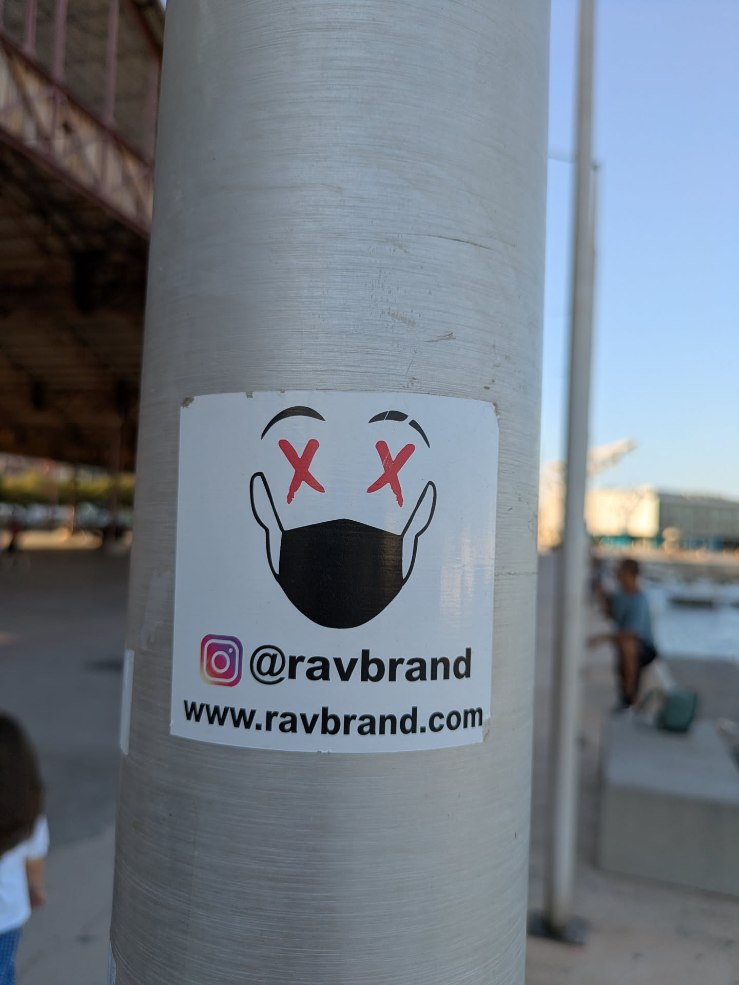 A sticker adhered to a pole. The sticker depicts a face with X's in place of eyes, and a mask covering the mouth and nose. Below the image are social media handles and a website URL.