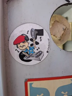 Circular sticker depicting a cartoon DJ with a red beret and Hawaiian shirt mixing on turntables. He holds a vinyl record aloft amidst floating music notes. An Instagram handle is printed on the side.