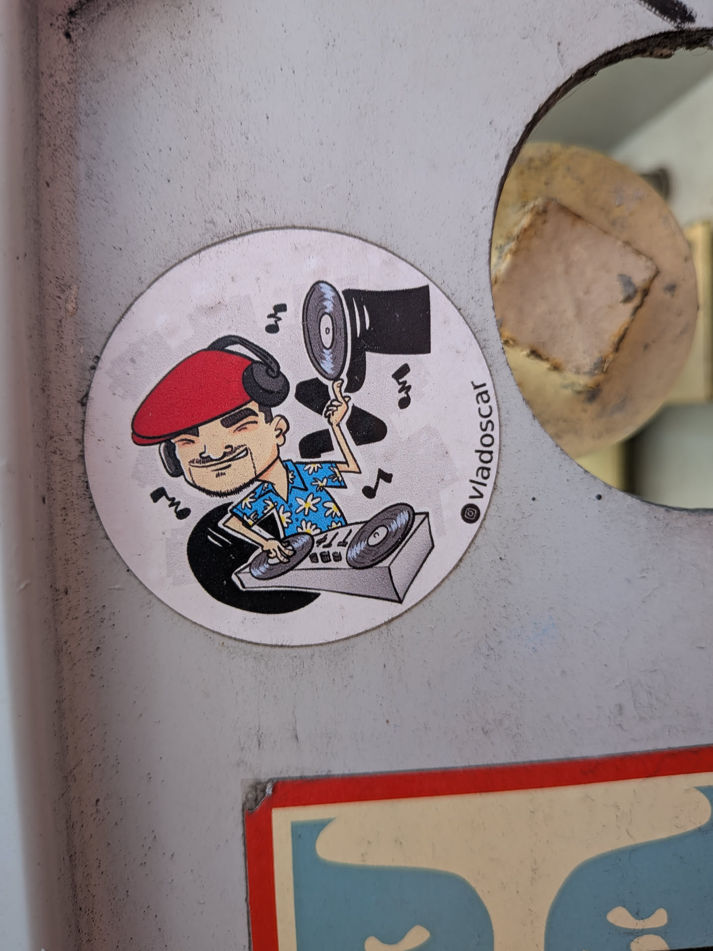Circular sticker depicting a cartoon DJ with a red beret and Hawaiian shirt mixing on turntables. He holds a vinyl record aloft amidst floating music notes. An Instagram handle is printed on the side.