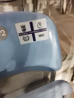 Street sticker A rectangular white sticker featuring a prominent dark blue cross. The sticker is divided into quadrants by the cross. In the top left quadrant, there is a black and white crest depicting a crowned figure. The top right quadrant displays a stylized intertwined 'C' and 'F' with a crown. The bottom left quadrant shows another black and white crest, possibly a 'C' and 'F' within a wreath and crown. The horizontal bar of the cross contains the text "MADRID FOOTBALL CLUB" in dark blue capital letters. The bottom right quadrant shows the year "1902" in dark blue numerals. The sticker has a slightly glossy finish and is applied to a light blue stadium seat.