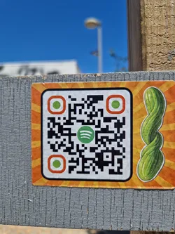 Street sticker A sticker featuring a QR code that links to a Spotify playlist. The sticker also has an illustration of a pickle on it. The background is orange with a sunburst design. The QR code has a green Spotify logo in the center.