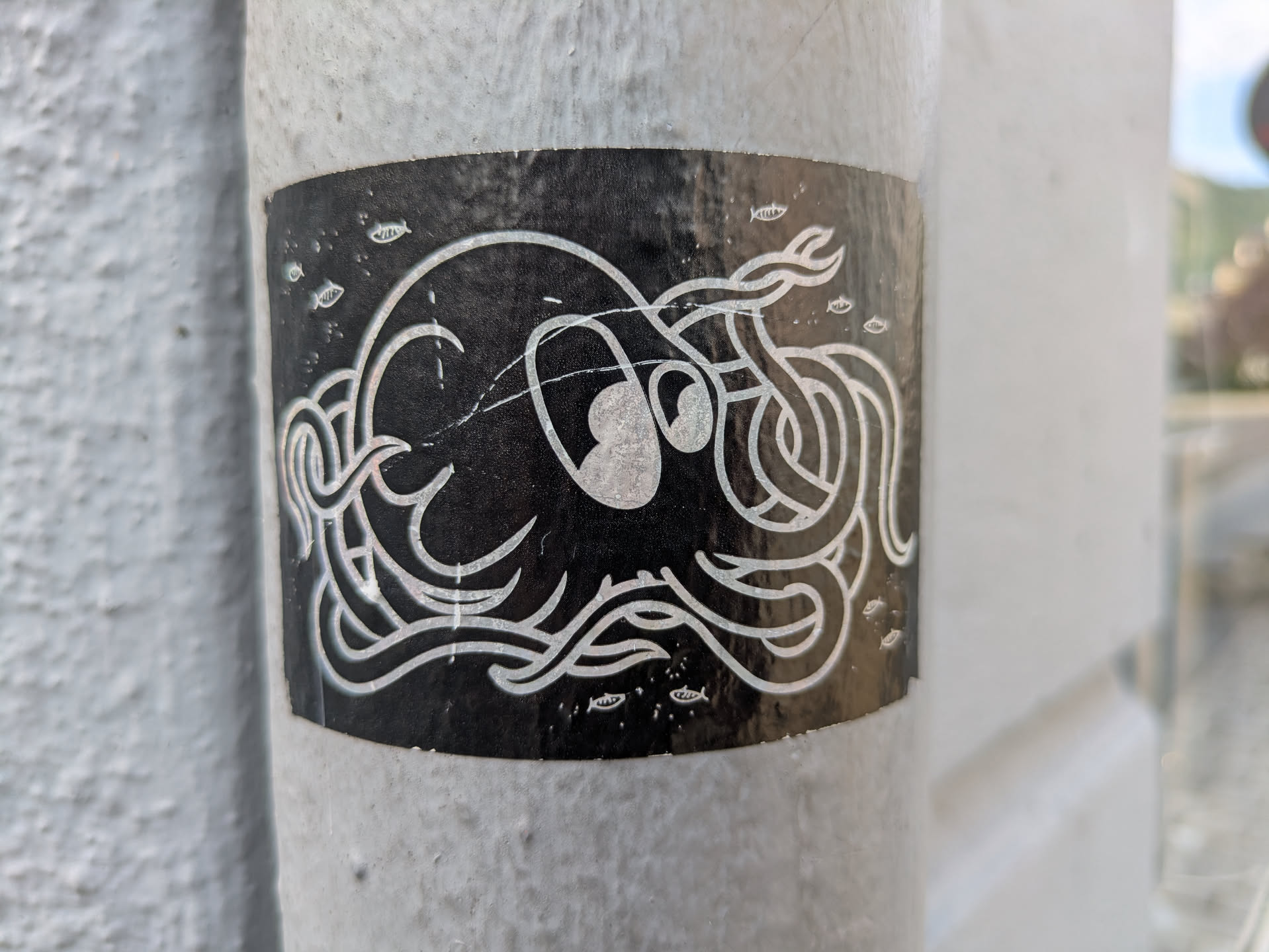 The sticker depicts an abstract, stylized octopus or kraken-like sea creature with swirling tentacles, rendered in light-colored (possibly white or silver) lines against a solid black background. Several small, simple fish shapes are scattered around the main creature. The sticker is placed on a textured, light-colored pole or column, showing some signs of wear and scratches on its surface.